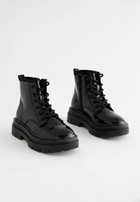 Bottines en cuir verni noir avec un laçage à l'avant et des semelles en caoutchouc épaisses. Les caractéristiques notables incluent une doublure texturée et une boucle de traction.