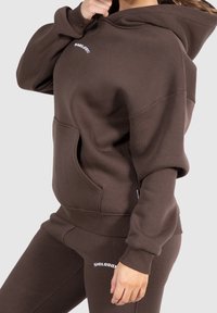 Felpa marrone con tasca frontale, polsini e orlo a coste. Presenta un logo bianco sul petto. Abbinata a pantaloni marroni coordinati, anch'essi con logo.