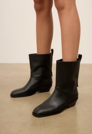 Benen met zwarte leren laarzen tot halverwege de kuit met lage schuine hakken, staand op een gladde beige vloer tegen een effen muur.