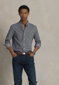 Grå skjorte med knapper laget av bomull, sammen med mørk denimjeans. Bæreren har en atletisk bygning, hendene i lommene, og et klokke på håndleddet.