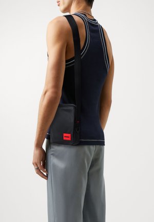Borsa a spalla nera con etichetta rossa, caratterizzata da un design rettangolare elegante e una tracolla regolabile, indossata sopra una canotta blu navy.
