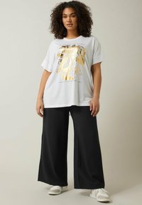 Witte t-shirt met een gouden bloemenpatroon en tekst, gecombineerd met zwarte wijde broek en witte sneakers. Glad materiaal, relaxed fit.