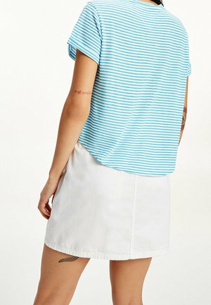 Top a maniche corte blu chiaro e bianco a righe con una vestibilità rilassata, abbinato a una gonna di jeans bianca. Tatuaggio evidente sul braccio.