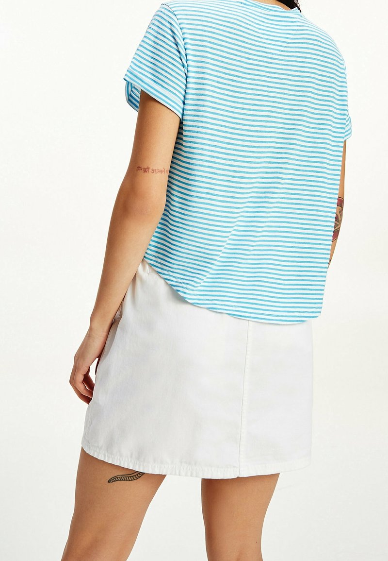 Svetle modré a bílé pruhované tričko s krátkým rukávem, uvolněného střihu, spárované s bílou denimovou sukní. Pozoruhodné tetování na paži.