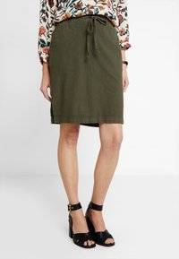 Jupe en coton vert olive avec un waistband à cordon, de longueur genou, présentant des fentes latérales et un design d'ourlet simple. Portée avec des sandales à talons noires.