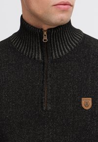Zwarte gebreide trui met een hoge geribbelde kraag, halflange rits en een bruine leren logo label met een berenmotief. Textuur van de stof.