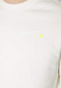 Bílá mikina vyrobená z měkké tkaniny se bílým logem Nike a malým žlutým srdíčkem poblíž loga, kulatým výstřihem a žebrovanými manžetami.