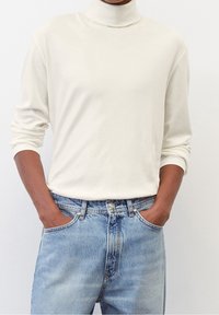 Langärmliges creamfarbenes Rollkragenoberteil, kombiniert mit hellblauen Denim-Jeans. Das Oberteil hat eine weiche, glatte Textur und einen entspannten Schnitt.