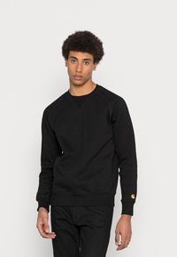 Felpa nera con collo rotondo, maniche raglan, polsini a coste e un accento dorato del logo sulla manica sinistra. Tessuto liscio e strutturato.