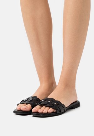 Feet wearing black flat slide sandals with woven straps and small silver studs on a white background.
