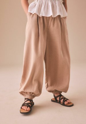Kind trägt lockere beige Hosen mit elastischen Bündchen und schwarze Riemchensandalen, steht vor neutralem Hintergrund.