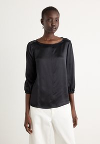 Zwart satijnen blouse met driekwart pofmouwen, ronde halslijn, gladde textuur en een iets relaxte pasvorm. Gedragen met lichtgekleurde broeken.