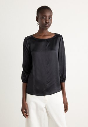 Blusa di raso nera con maniche a sbuffo a tre quarti, scollatura rotonda, texture liscia e una vestibilità leggermente rilassata. Indossata con pantaloni di colore chiaro.