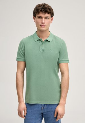 Giovane uomo con capelli corti e ricci che indossa una polo verde chiaro e jeans blu, in piedi davanti a uno sfondo uniforme.