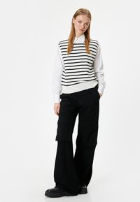 Chaleco de punto a rayas blancas y negras sobre una camisa blanca con cuello, combinado con pantalones cargo anchos negros y calzado negro de gran tamaño.
