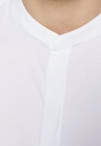 Biała koszula z okrągłym kołnierzykiem, wyposażona w ukrytą listwę na guziki oraz gładką, odporną na zagniecenia strukturę. Zbliżenie na dekolt.