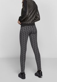 Černá bomber bunda s vzorovaným zlatým logem, v kombinaci s těsnými černými legínami s opakovaným potiskem loga. Černá obuv celý look dotváří.