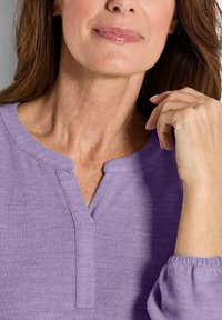 Lila Henley-Shirt mit V-Ausschnitt, gerippter Textur und langen Ärmeln. Leichter Stoff zeigt sanfte Falten und einen natürlichen Fall.