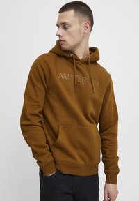 Sweat à capuche marron avec poche kangourou, cordon de serrage au niveau de la capuche, et "AMSTERDAM" brodé en couleur claire sur la poitrine. Texture de tissu douce.