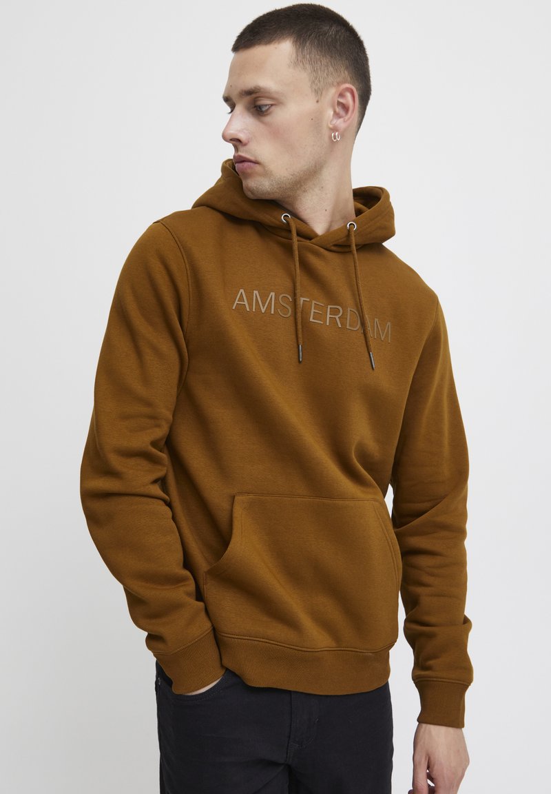 Sweat à capuche marron avec poche kangourou, cordon de serrage au niveau de la capuche, et "AMSTERDAM" brodé en couleur claire sur la poitrine. Texture de tissu douce.