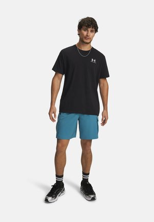 Zwart T-shirt met korte mouwen en een wit logo, gecombineerd met turquoise korte broek met zijzakken en zwarte sportschoenen met zilveren accenten.