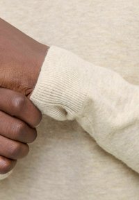 Beige Pullover-Detail mit geripptem Bund, weicher Textur und sichtbaren Nähten. Hand hält den Bund vor einem neutralen Hintergrund.