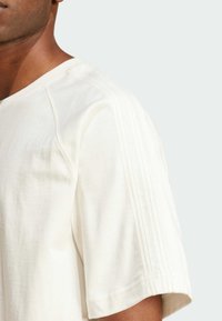 Närbild av en person som bär en vit kortärmad skjorta med subtila vertikala sömdetaljer längs axeln och ärmen.