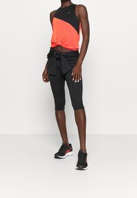 Frau in einem ärmellosen Oberteil in Schwarz und Orange, schwarzen cropped Leggings, schwarzer Jacke um die Taille gebunden und schwarzen Laufschuhen mit orangenen Akzenten.