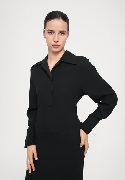 Bluză lungă cu mâneci negre, cu guler, având o deschidere fără nasturi, textură netedă și o siluetă ajustată. Design simplu și minimalist.