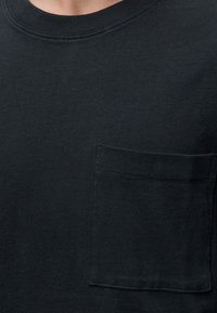 Κοντινό πλάνο μαύρου βαμβακερού T-shirt με στρογγυλή λαιμόκοψη και τσέπη στο στήθος στην αριστερή πλευρά.