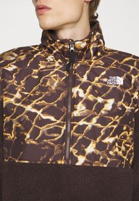 Zbliżenie ujęcia osoby noszącej brązową polarową kurtkę z żółtym marmurkowym wzorem i zamkiem błyskawicznym, z logo The North Face na piersi.