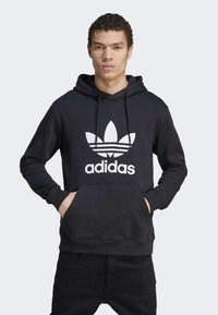 Crna pamučna dukserica s kenguruh džepom, kapuljačom s uže, i velikim bijelim Adidas logom koji sadrži tri pruge na dizajnu lista.