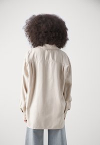 Dalla camicia a righe beige con colletto, maniche lunghe e polsini abbottonati. Il tessuto sembra liscio e leggero.