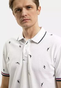 Biała koszulka polo z teksturowanej tkaniny, ozdobiona małą czarną haftowaną aplikacją w motywie sportowym. Wykończona granatowymi i czerwonymi paskami przy kołnierzyku.