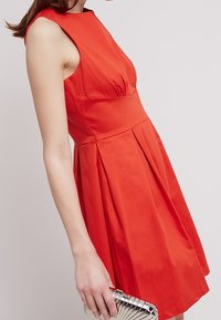 Donna in un vestito rosso senza maniche tiene una piccola borsa a pochette metallica argento contro uno sfondo chiaro e semplice.