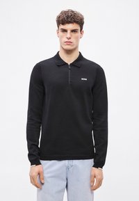 Črna polo majica z dolgimi rokavi z zadrgo na ovratniku, žametno strukturo in majhnim belim logotipom na prsih, kombinirana s svetlo modrimi kavbojkami.