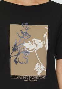T-shirt noir avec un graphique carré beige orné de motifs floraux blancs et pailletés en bleu, accompagné du texte « ÉLEVÉ CHAQUE JOUR classique moderne. »