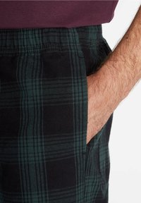 Pantalon plissé avec motif à carreaux noirs et verts, doté d'une ceinture élastique et d'une poche latérale. Le tissu semble doux et texturé.