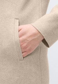 Μπεζ παλτό από τσόχα με πλαϊνή τσέπη. Κοντινό πλάνο χεριού τοποθετημένου μέσα στην τσέπη, που αναδεικνύει την υφή του υφάσματος και τη λεπτομέρεια της ραφής.