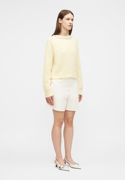 Γυναίκα με ίσια καστανά μαλλιά που φοράει χνουδωτό πουλόβερ σε ανοιχτό κίτρινο χρώμα, λευκά σορτς μέχρι το γόνατο και λευκά παπούτσια slingback με χαμηλό τακούνι.