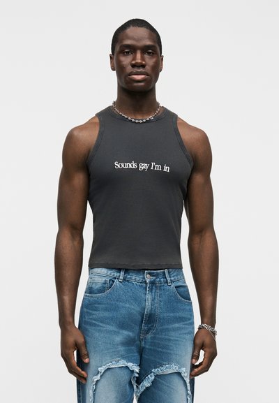 Σκούρο γκρι ριμπ crop top με λευκό κείμενο, συνδυασμένο με μπλε τζιν παντελόνι με σκισίματα. Το μοντέλο φοράει ασημένιο κολιέ με αλυσίδα. Απλή φόντο.