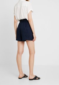 Námorní modré kraťasy s uvolněným střihem, vybavené elastickým pasem. Spárované s volnou bílou košilí a černými klouzavými sandály s akcenty.