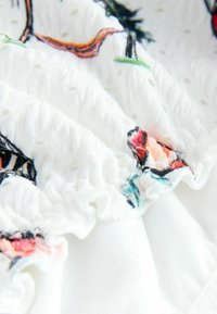 Tăietură din bumbac alb cu finisaj texturat, având broderie florală colorată în roz, roșu și verde. Detalii cu margini volan.