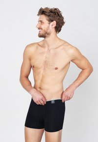 Zwarte boxershorts met een gladde textuur, voorzien van een grijze tailleband met een logo, gedragen door een mannelijke model. Geen andere kleding of accessoires.