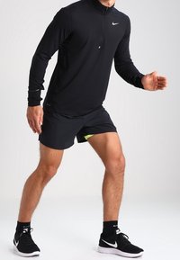 Schwarzes, langärmliges Sportoberteil mit Reißverschlusskragen, kombiniert mit schwarzen Shorts mit neongrünen Akzenten. Schwarze Sportschuhe runden den Look ab.