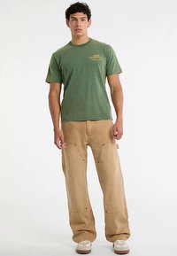 Groen katoenen T-shirt met geel logo, gecombineerd met beige baggy cargobroek met meerdere zakken en een losse pasvorm, gedragen met beige sneakers.