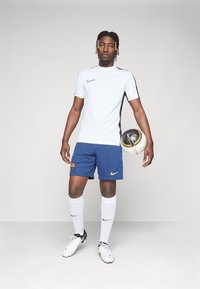 Balta Nike t-krekls ar melniem sānu akcentiem, zili šorti ar Barselonas logo, balti zeķes un melni futbols čības. Tur rokās futbolu.
