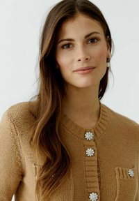 Brauner Strickcardigan mit Rundhalsausschnitt, ausgestattet mit vier dekorativen blumenförmigen Knöpfen und einer Fronttasche. Glatte Textur.