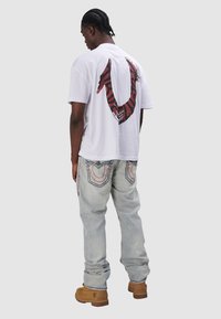 Biała koszulka z dużą czerwoną grafiką na plecach, w połączeniu z jasnoniebieskimi dżinsami z haftowanymi akcentami na kieszeniach i brązowymi butami.