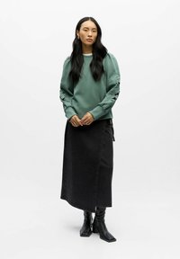 Sweatshirt vert à manches bouffantes avec découpes florales, associé à une jupe maxi en denim noir et des bottes noires à bout carré.